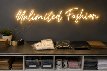 A glowing neon sign reading 'Unlimited Fashion' mounted on a dark gray wall above a counter displaying folded clothing, packaging, and storage bins in a retail or boutique setting.